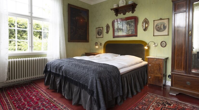 Ett fönster, en säng, ett klädskåp i ett rum med grön tapet och många tavlor.
